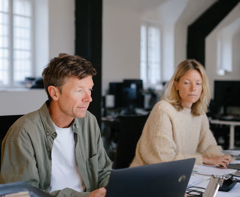 Man and woman working on laptops in a modern office with large windows and black beams.