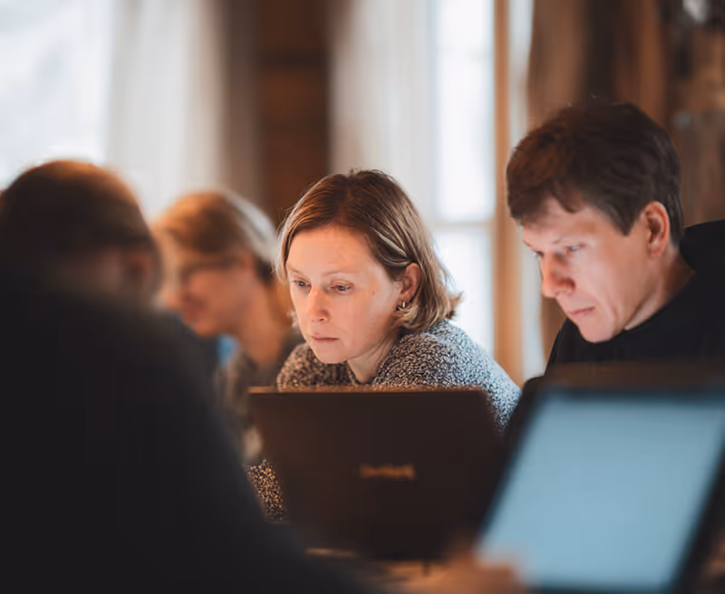 Group of focused young adults working on laptops in a warmly lit room with large windows.