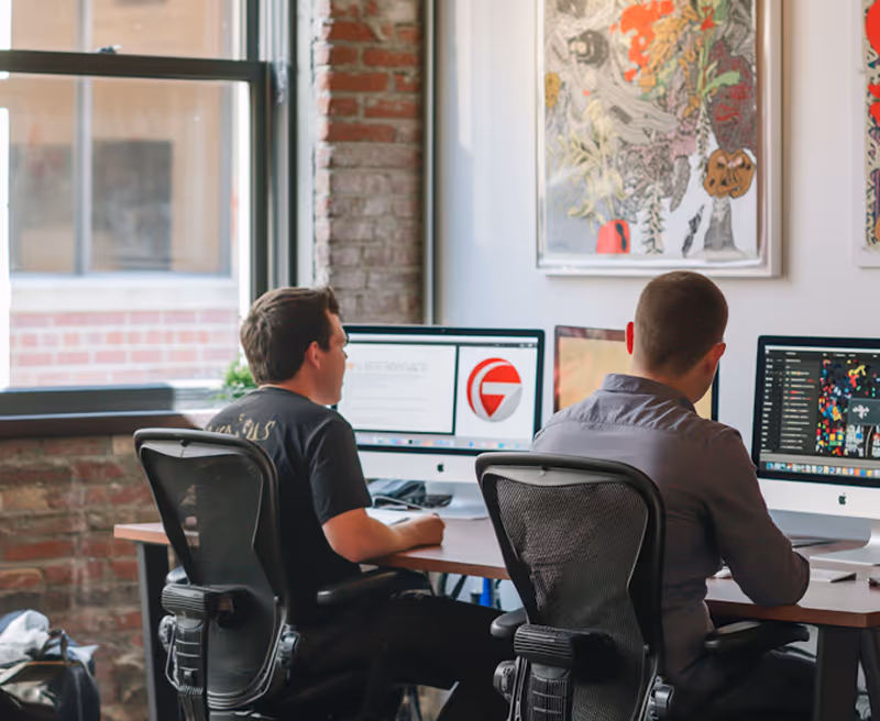 Two men working on desktop computers in a modern office with brick walls and framed artwork.